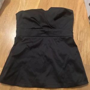 Svart tubtopp från Pimkie - Säljer en svart axelbandslös tubtopp från Pimkie. Toppen har en markerad midja och lätt utsvängd nederdel. Sitter uppe med hjälp av silikonband på insidan och har en slät, stilren look. Perfekt för dig som gillar minimalistisk stil. Liten defekt see i bild 4