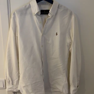 Vit skjorta från Ralph Lauren Slim Fit - Klassisk vit skjorta från Ralph Lauren i slim fit-modell. Skjortan har lång ärm, knappar framtill och ikoniska broderade loggan på bröstet. Tillverkad i mjuk bomull och passar perfekt till en clean och stilren look. Skjortan är i sjukt bra skick!
