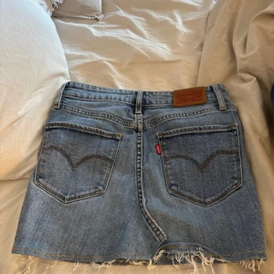Levi's jeanskjol med fransig kant - Säljer en klassisk blå jeanskjol från Levi's med hög midja och fransig rå kant nertill. Kjolen har fem fickor, bälteshällor och Levi's ikoniska läderpatch bak. 
