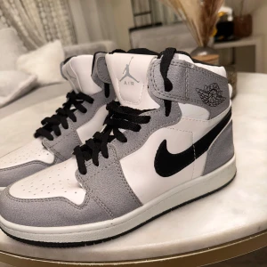Jordan 1 Smoke grey - Snygga Nike Air Jordan 1 High sneakers i vitt och grått med svarta detaljer och klassisk svart swoosh. Ovandel i skinn och mocka, svarta skosnören och ikonisk Air Jordan-logga på sidan. Perfekta för dig som vill sticka ut med en tidlös streetstil.