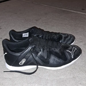 Svarta fotbollsskor från Puma strl 41 - Svarta fotbollsskor från Puma med vita detaljer och logga på sidan och hälen. Skorna har snörning och en lätt, syntetisk ovandel med prickigt mönster. Sulan är vit och designen är sportig och modern, perfekt för dig som gillar att sticka ut på planen.