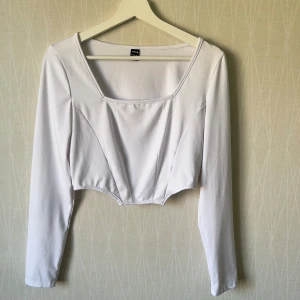 Vit croppad långärmad topp - Säljer en vit croppad topp  med lång ärm och fyrkantig ringning. Toppen har markerade sömmar framtill som ger en lätt korsettkänsla och är tillverkad i ett stretchigt, mjukt material. Perfekt för dig som gillar stilrena och trendiga plagg.