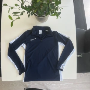 Nike marinblå långärmad träningströja - Nike långärmad träningströja i marinblått med vita detaljer längs ärmarna och sidorna. Tröjan har en halv dragkedja framtill och klassisk Nike-logga på bröstet. Perfekt för sport eller chill dagar. Tillverkad i mjukt och lätt polyestermaterial.