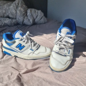 New Balance 550 vita och blå sneakers - Snygga New Balance 550 sneakers i vitt och blått med klassisk retrostil. Skorna har blå detaljer, vit ovandel i skinn och mesh, samt tydlig logga på sidan. Lågt skaft, snörning och platt sula gör dem perfekta för en sportig look.