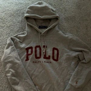 Grå hoodie från Polo Ralph Lauren - Snygg grå hoodie från Polo Ralph Lauren med stor röd POLO-text på bröstet och broderad logga på ärmen. Klassisk huva med snörning och mjukt bomullsmaterial. Perfekt för en chill och stilren look.
