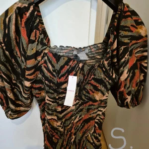 Mönstrad kortklänning från Lindex - Cool kortklänning från Lindex med tigerinspirerat mönster i svart, orange, beige och grönt. Klänningen har puffiga korta ärmar och smockad överdel som ger en snygg passform. Perfekt för dig som vill sticka ut med en färgstark look.