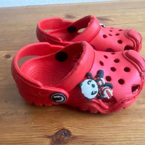 Röda barn  sandaler med panda-dekoration - Coola röda sandaler från Deluxe med ventilerande hål och en söt panda-dekoration framtill. Skorna har en justerbar hälrem och är tillverkade i lätt syntetmaterial, perfekta för chill dagar eller bad. Sulan är rejält mönstrad för extra grepp.