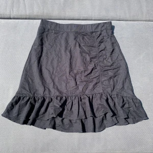 Svart volangkjol med hög midja - Supersnygg svart kjol med volangkant nertill och hög midja. I sidan har kjolen en liten scrunch. Kjolen har en lätt A-linjeform och är perfekt för dig som gillar en stilren look med något lite extra. Passar till både sneakers och boots. Lite liten i storleken.