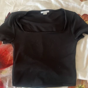 Svart topp med fyrkantig ringning Amisu - Säljer en svart, tight topp från Amisu med korta ärmar och snygg fyrkantig ringning. Toppens material är stretchigt och känns mjukt mot huden, perfekt att styla med jeans eller kjol. Enkel och stilren design som passar till många looks.