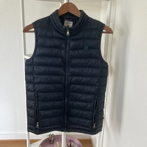 Mörkblå dunväst från U.S. Polo Assn. - Snygg mörkblå dunväst från U.S. Polo Assn. med quiltad design och dragkedja framtill. Västen har två praktiska fickor med dragkedja och en hög krage. Perfekt att slänga över en hoodie för en sportig look.