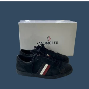Svarta sneakers från Moncler mocka - Stilrena svarta sneakers från Moncler i mocka med klassiska Moncler-ränder i vitt och rött på sidan. Skorna har snörning och rund tå, samt diskret Moncler-logga på sidan och plösen. Perfekta för dig som gillar exklusiva och sportiga vibes. Levereras med originalförpackning och dustbags. Allt og ingår! Kom med bud.