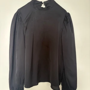 Svart blus med puffärm från Vila - Stilren svart blus från Vila i storlek 34. Blusen har långa puffärmar, hög hals med knappstängning bak och en lätt flowy passform. Perfekt att styla med jeans eller kjol för en clean look.