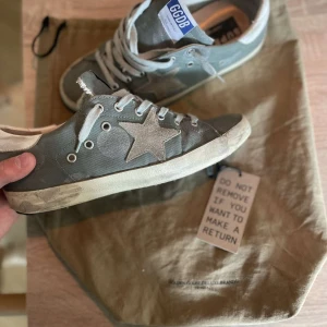 Golden Goose Superstar - Golden Goose Superstar sneakers i grått med vit stjärndetalj på sidan och vita snören. Skorna har en vintage-inspirerad look med slitna detaljer, rund tå och platt sula. Ovandelen är i canvas och detaljer i mocka. Perfekta för dig som gillar streetstyle med edge.