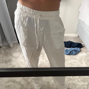 Vita linnebyxor med snörning - Säljer ett par vita linnebyxor med elastisk midja och snörning framtill. Byxorna har avslappnad passform och raka ben, perfekta för varma dagar. De har fickor på sidorna och är tillverkade i ett lätt och luftigt linnematerial.