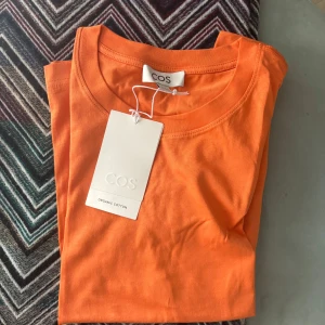 Orange t-shirt från COS i bomull - Snygg orange t-shirt från COS i ekologisk bomull. Klassisk rund halsringning och korta ärmar. Perfekt basic-plagg med clean look och skön passform. Enkel att matcha med jeans eller shorts för en chill stil.
