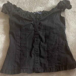 Svart offshoulder blus med volang - Supersöt svart blus från brandy Melville med offshoulder och volangkant. Knappar framtill, rynkade detaljer vid axlarna och smala veck längs framsidan. Väldigt bra skick
