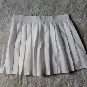 Vit plisserad kjol från Gina Tricot - Superfin vit plisserad kjol från Gina Tricot med bred resår i midjan. Kjolen är kort och har en klassisk tennisstil som är perfekt för en trendig look. Materialet är mjukt och lätt, vilket gör den bekväm att bära hela dagen.