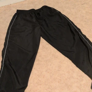 Svarta mjukisbyxor från Units - Svarta mjukisbyxor från Units med elastisk midja och tunna vita ränder längs sidorna. Byxorna har en avslappnad passform och är tillverkade i ett mjukt syntetmaterial som känns skönt mot huden.