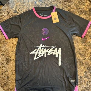 PSG x Stüssy x Nike svart fotbollströja - Svart PSG x Stüssy x Nike fotbollströja i Dri-FIT-material. Tröjan har korta ärmar, rosa detaljer vid hals och ärmslut, samt diskret blommönster i tyget. Stüssy-logga i vitt framtill och PSG-emblem i lila. Perfekt för dig som gillar streetwear och fotboll.