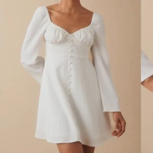 Vit kortklänning med knappar - Superfin vit kortklänning med långa ärmar och markerad byst. Klänningen har dekorativa knappar framtill och en lätt utsvängd kjol. Perfekt för sommarens alla tillfällen och ger en romantisk vibe.
