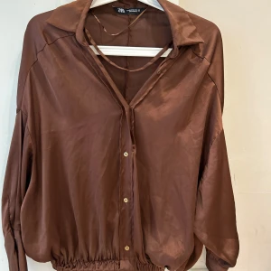 Brun satinblus från Zara - Snygg brun blus från Zara i glansigt satinliknande material. Modellen har v-ringning, krage, knappar framtill och resår i midjan för en avslappnad men chic look. Långa ärmar med breda manschetter och en lös passform.