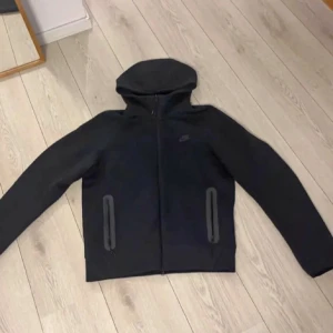 Svart Nike vindjacka med huva - Svart vindjacka från Nike med hel dragkedja och två praktiska fickor framtill. Jackan har en stilren look med huva och diskret Nike-logga på bröstet. Perfekt för dig som gillar sportig och clean stil.