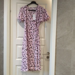 Lilarosa omlottklänning från Vila - Söt lila omlottklänning från Vila med korta ärmar och svart/vitt blommigt mönster. Klänningen har v-ringning och knyt i midjan för en snygg passform. Perfekt för dig som gillar en feminin och trendig stil.