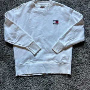 Vit sweatshirt från Tommy Jeans - Vit sweatshirt från Tommy Jeans med broderad logga på bröstet. Klassisk rund halsringning, ribbade muddar och avslut. Tillverkad i mjuk bomull, perfekt för en clean och stilren look.