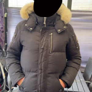 Svart dunjacka från Moose Knuckles - Säljer en svart dunjacka från Moose Knuckles med fluffig beige pälskrage. Jackan har flera dragkedjor, hög krage och logga på ärmen. Perfekt för kalla dagar med sin tjocka vaddering och snygga detaljer. Klassisk och stilren modell med lång ärm och praktiska fickor.