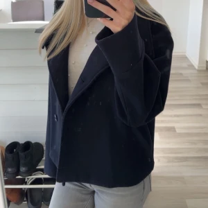 Kort kappa  - Snygg mörkblå kappa i ullmix med rak, boxig passform och hög krage. Jackan har stora knappar framtill och vida ärmar för en chill vibe. Perfekt till jeans eller kostymbyxor för en clean look.