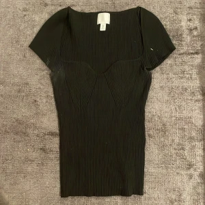 Svart ribbad topp från H&M - Svart ribbad topp från H&M med korta ärmar och figurnära passform. Toppen har en snygg v-ringad skärning och markerade sömmar över bysten som ger en cool struktur. Perfekt att styla med jeans eller kjol för en trendig look.
