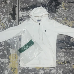 Vit hoodie från Polo Ralph Lauren - Vit hoodie från Polo Ralph Lauren med dragkedja och huva. Klassisk design med broderad logga på bröstet och fickor framtill. Perfekt för en chill och stilren look. Mjuk bomullskänsla och snygga detaljer som snörning i huvan.