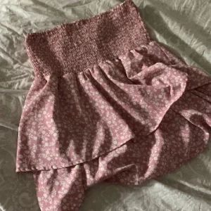 Rosa blommig volangkjol - Supersöt rosa kortkjol med små vita blommor och bred smock i midjan. Kjolen har volang och omlottkänsla, perfekt för en somrig look. Lätt och luftig, passar till sneakers eller sandaler.