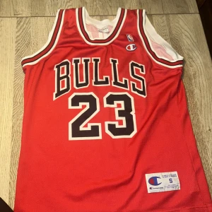 Chicago Bulls Jordan 23 basketlinne - Säljer ett rött Chicago Bulls basketlinne från Champion med nummer 23 och Jordan-tryck på ryggen. Klassisk NBA-stil med vita och svarta detaljer runt ärm och hals. Luftigt meshmaterial, perfekt för basket eller streetwear.