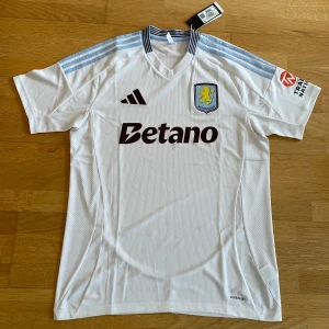 Aston Villa bortatröja Adidas L - Aston Villa bortatröja från Adidas i vitt med ljusblå detaljer på axlarna och klubbmärke på bröstet. Tröjan har korta ärmar, ribbad struktur och Aeroready-material som andas. Snygg krage med blå och vita ränder samt sponsortryck på bröst och ärm.