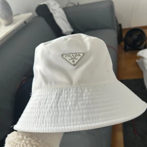 Vit bucket hat från Prada - Snygg vit bucket hat från Prada med klassisk triangellogo framtill. Hatten är tillverkad i glansig syntet och har en stilren, minimalistisk design. Perfekt accessoar för att lyfta din outfit med en exklusiv touch.