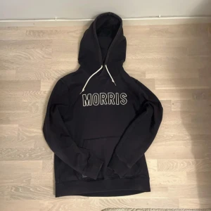 Svart hoodie från Morris med tryck - Svart hoodie från Morris med vit broderad logga på bröstet och vita snören i huvan. Klassisk känguruficka framtill och långärmad modell. Perfekt för en avslappnad och stilren look. Pris kan diskuteras vid snabb affär