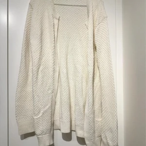 Vit stickad kofta med struktur - Mysig vit stickad kofta med snygg struktur och ribbade muddar. Koftan är öppen framtill och har långa ärmar, perfekt att slänga på sig när det är lite kyligt. Enkel att matcha med olika outfits och superbekväm att bära.