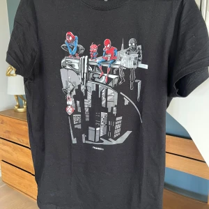 Ny Svart Spiderman t-shirt från Friking S - Svart t-shirt från Friking med coolt Spiderman-tryck där flera olika Spiderman-karaktärer sitter på en balk ovanför en stad. T-shirten har korta ärmar och är gjord i mjuk bomull. Perfekt för dig som gillar Marvel och vill sticka ut med en unik design. Oanvänd 