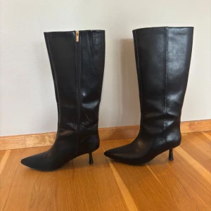 Svarta knähöga boots från ASOS - Knappt använda. Stilrena svarta knähöga boots från Asos med spetsig tå och smal klack. Skorna har dragkedja på insidan och är tillverkade i syntetmaterial med en slät finish. Ursprungligt pris 682kr. 6cm hög klack.