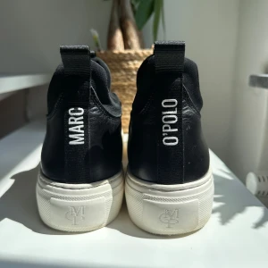 Svarta sneakers från Marc O'Polo - Stilrena och bekväma slip-on sneakers från Marco Polo i helsvart design. Skorna har en modern och minimalistisk look utan snörning (lace-less), vilket gör dem enkla att ta på och av – perfekta för både vardag och mer uppklädda tillfällen. De karaktäristiska Marco Polo-detaljerna ger en diskret touch av kvalitet och design. Skorna är i bra skick med inga större tecken på slitage. Inga hål, defekter eller andra skador – endast lätt använda och väl omhändertagna.  