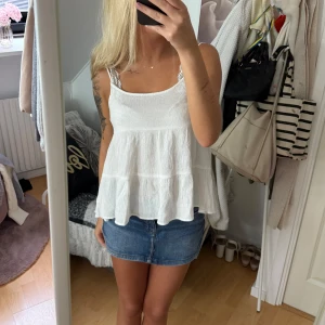 Vit volanglinne  - Supersöt vit linnetopp med tunna axelband och volangig, flowy passform. Toppen har en lätt skrynklig struktur och är perfekt för varma dagar. Enkel och stilren design som är lätt att matcha med jeans eller kjol.