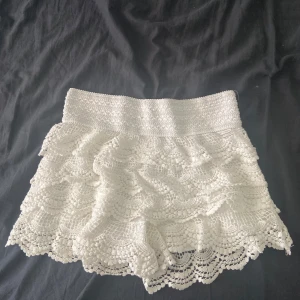 Vita virkade spets-shorts - Superfina vita shorts med bred resår i midjan och flera lager volangig virkad spets. De har en romantisk och bohemisk vibe, perfekta för varma dagar. Materialet är mjukt och luftigt, och spetsdetaljerna gör dem extra unika och söta.