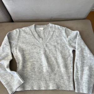 Grå stickad v-ringad tröja Gina Tricot - Mysig grå stickad tröja från Gina Tricot med v-ringning och breda ribbade muddar vid ärmslut och nederkant. Perfekt för lager-på-lager och chill dagar. Enkel att matcha med jeans eller kjol för en avslappnad look.