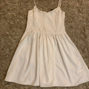 Vit spetsklänning från Gina Tricot - Superfin vit kortklänning från Gina Tricot med smala axelband och blommig spets upptill. Kjolen är lätt och luftig, perfekt för sommarens alla tillfällen. Dragkedja i ryggen för enkel påklädning och snygg passform.