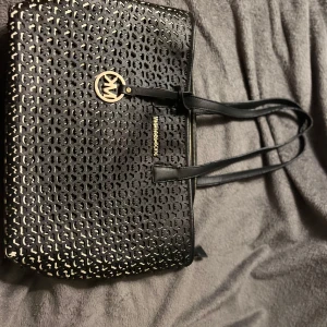 Svart shoppingväska Michael Kors - Snygg svart shoppingväska från Michael Kors med guldiga detaljer och MK-logga. Väskan har ett coolt perforerat mönster på framsidan och dubbla axelband. Tillverkad i skinnliknande material och stängs med dragkedja. Perfekt för dig som vill ha plats för allt du behöver. Älskat väskan så mycket men har köpt en nyare modell. Finns lite slit på axelbanden och dragkedjan stängs inte helt 1/10 gånger 