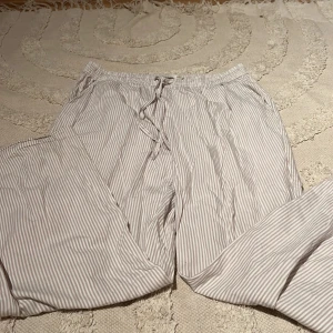 Randiga byxor från 157 med snörning - Luftiga byxor från 157 med beige och vita smala ränder. De har elastisk midja med snörning och är tillverkade i ett mjukt bomullstyg. Perfekta för chill dagar eller när du vill ha något bekvämt på dig