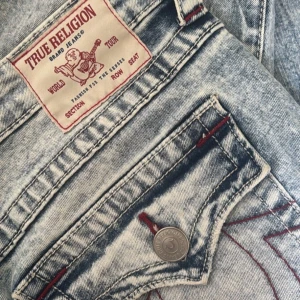 True Religion ljusblå bootcut jeans - Säljer ett par ikoniska ljusblå jeans från True Religion med bootcut passform. Jeansen har slitna detaljer på benen, klassiska bakfickor med lock och röda sömmar. Grov tvättad denim med snygga kontraster och silverfärgade knappar. Skriv privat för frågor. Använda ca 2 gånger💕