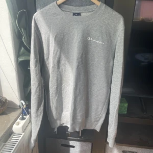 Grå sweatshirt från Champion M - Grå sweatshirt från Champion i storlek M. Klassisk crewneck med broderad Champion-logga på bröstet och liten logga på ärmen. Mjuk bomullsblandning och ribbade muddar vid ärmslut och midja. Perfekt för en chill och stilren look.