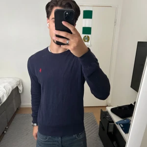 Mörkblå tröja från Ralph Lauren - Säljer en mörkblå långärmad tröja från Ralph Lauren med den klassiska röda loggan broderad på bröstet.  Är tillverkad i mjuk bomull. Storlek S slimfit.